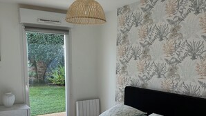 Een slaapkamer met een bed, nachtkastje, lamp, wand met bloemenprint en uitzicht op een tuin via een schuifdeur.