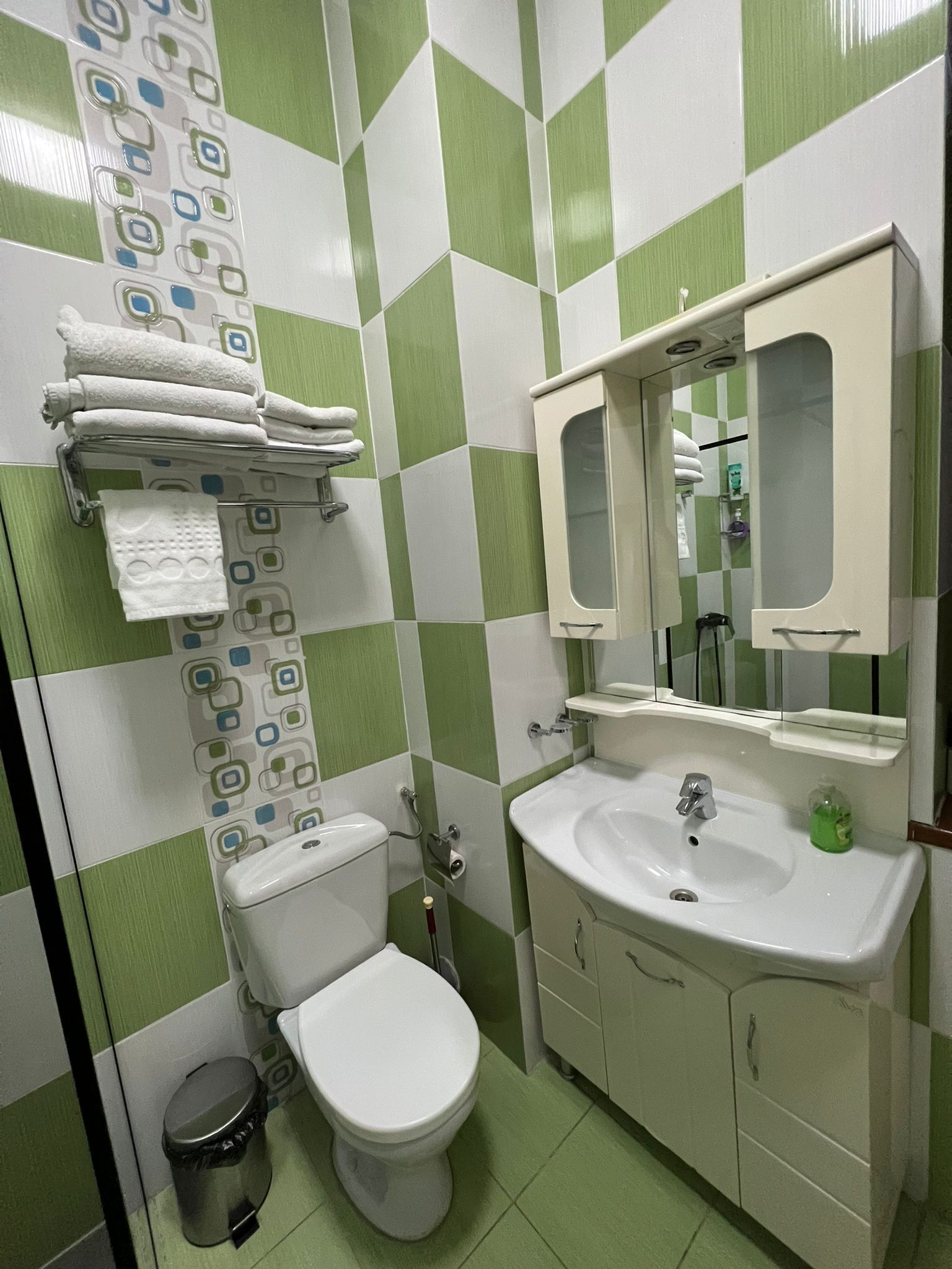 Un baño con inodoro, lavamanos y espejo. Hay toallas colgadas en un perchero y un bote de basura.
