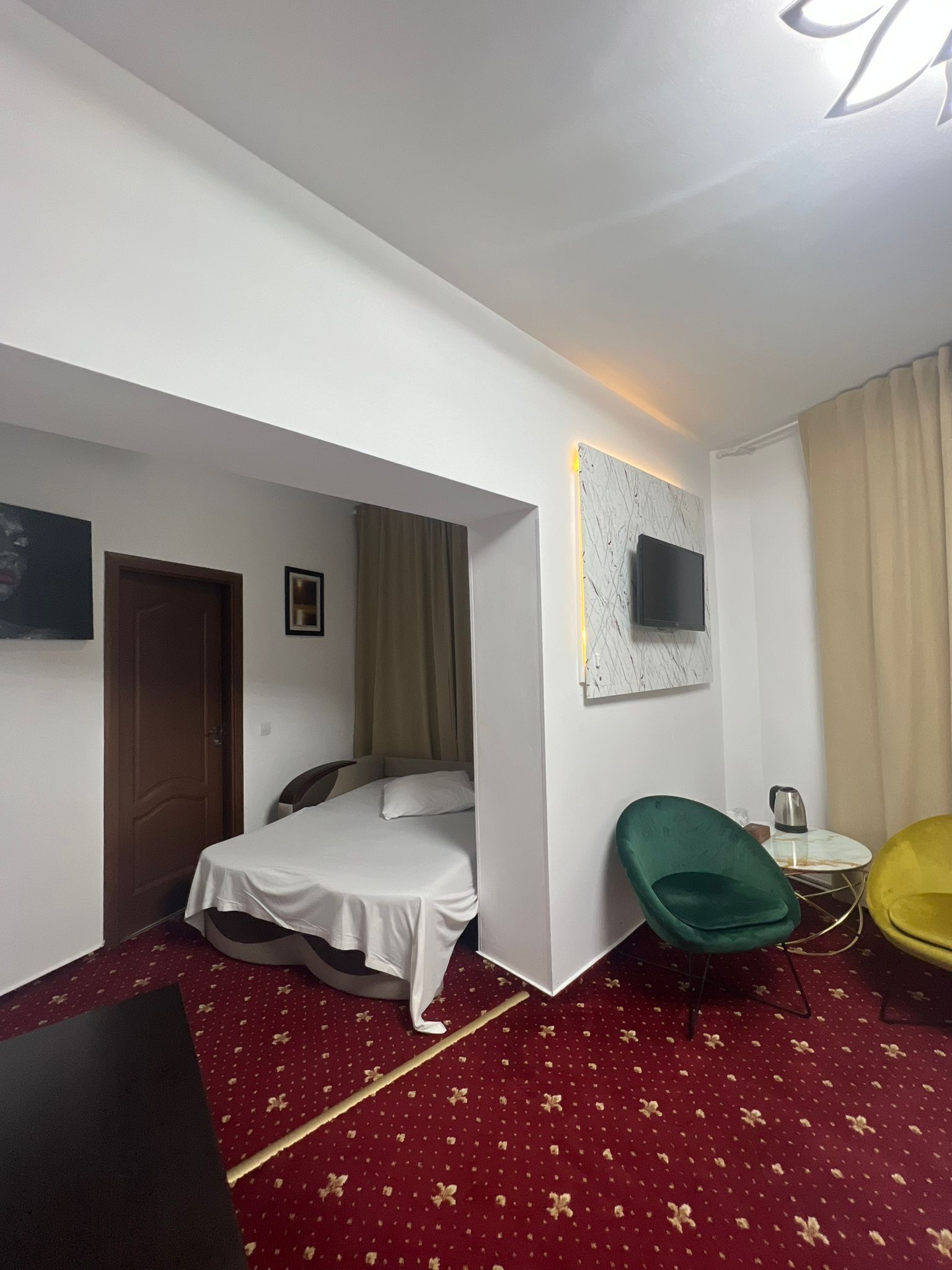 Une chambre d’hôtel avec un lit, un fauteuil vert, un fauteuil jaune, une petite table avec une bouilloire et un téléviseur fixé au mur.