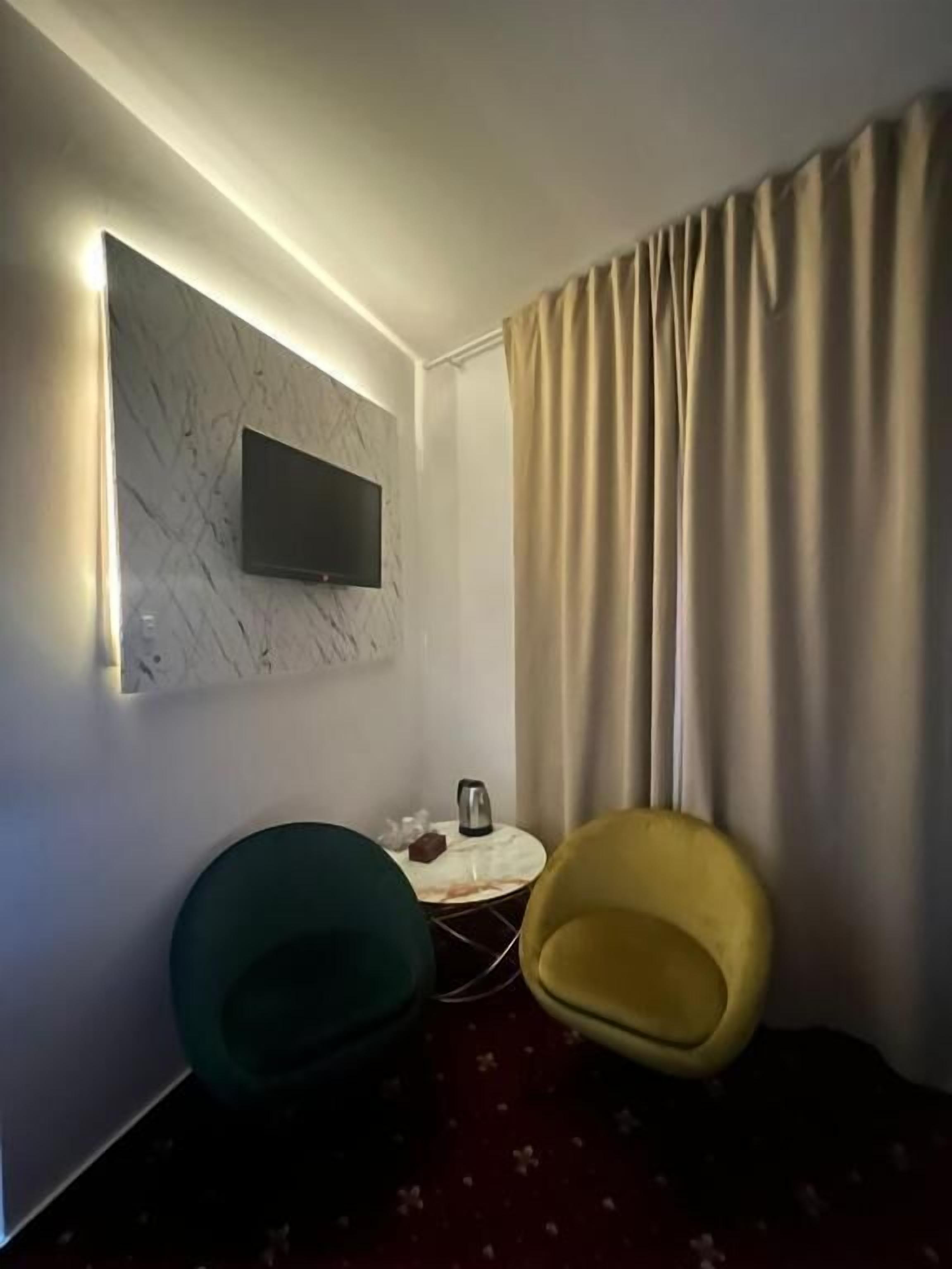 Una habitación moderna con un televisor de pantalla plana montado en una pared de mármol, una pequeña mesa redonda con una tetera y dos sillas, una verde y una amarilla.