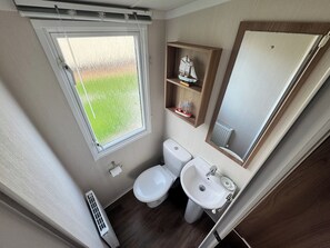 A compact bathroom with a window, a toilet, a sink, and a small shelf with decorative items. - The Pilots Rest Sleeps 6 (Exmouth)