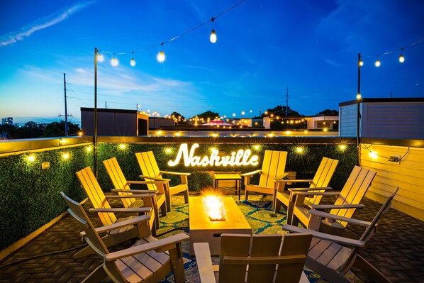 An outdoor seating area with Adirondack chairs and a fire feature, surrounded by string lights and a neon sign reading "Nashville".