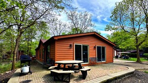 A small wooden cabin with a picnic table and grill, surrounded by trees and a paved patio. - Space to Laugh, Lounge & Live | 12 ppl | IC108 (Put-in-Bay)