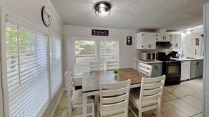 A kitchen with white cabinets, a microwave, and a stove. There is a dining table with chairs and a clock on the wall. - All New, All Comfort—Fits 12 ppl | IC110 (Put-in-Bay)