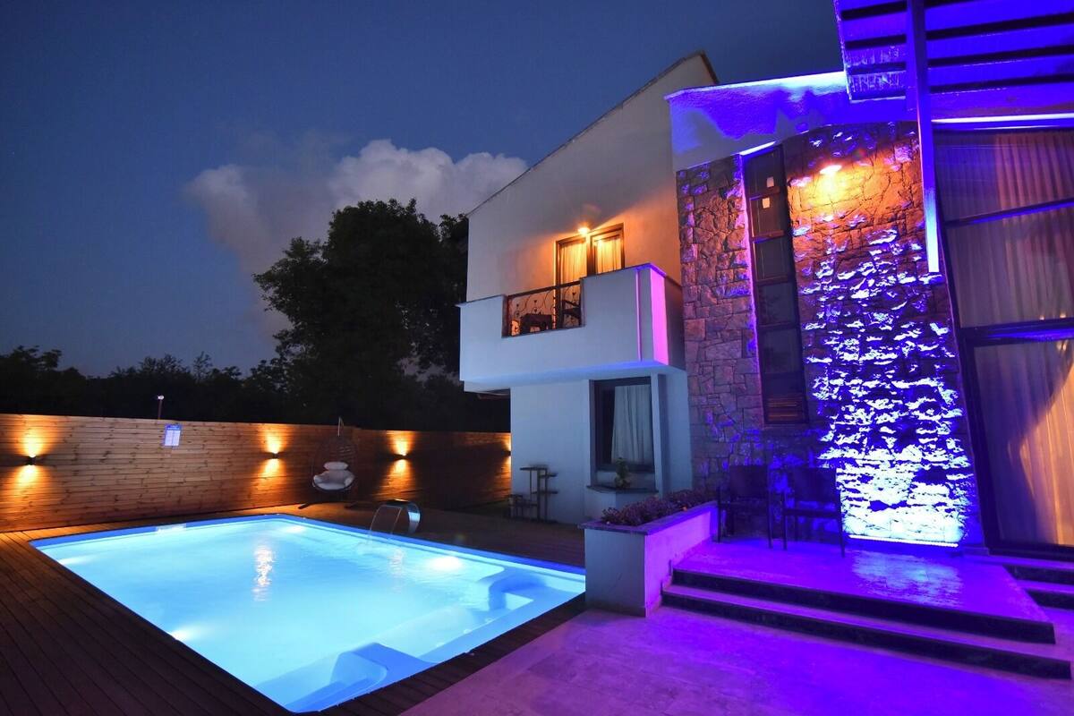 Una casa moderna con piscina, muro de piedra y balcón con iluminación exterior.