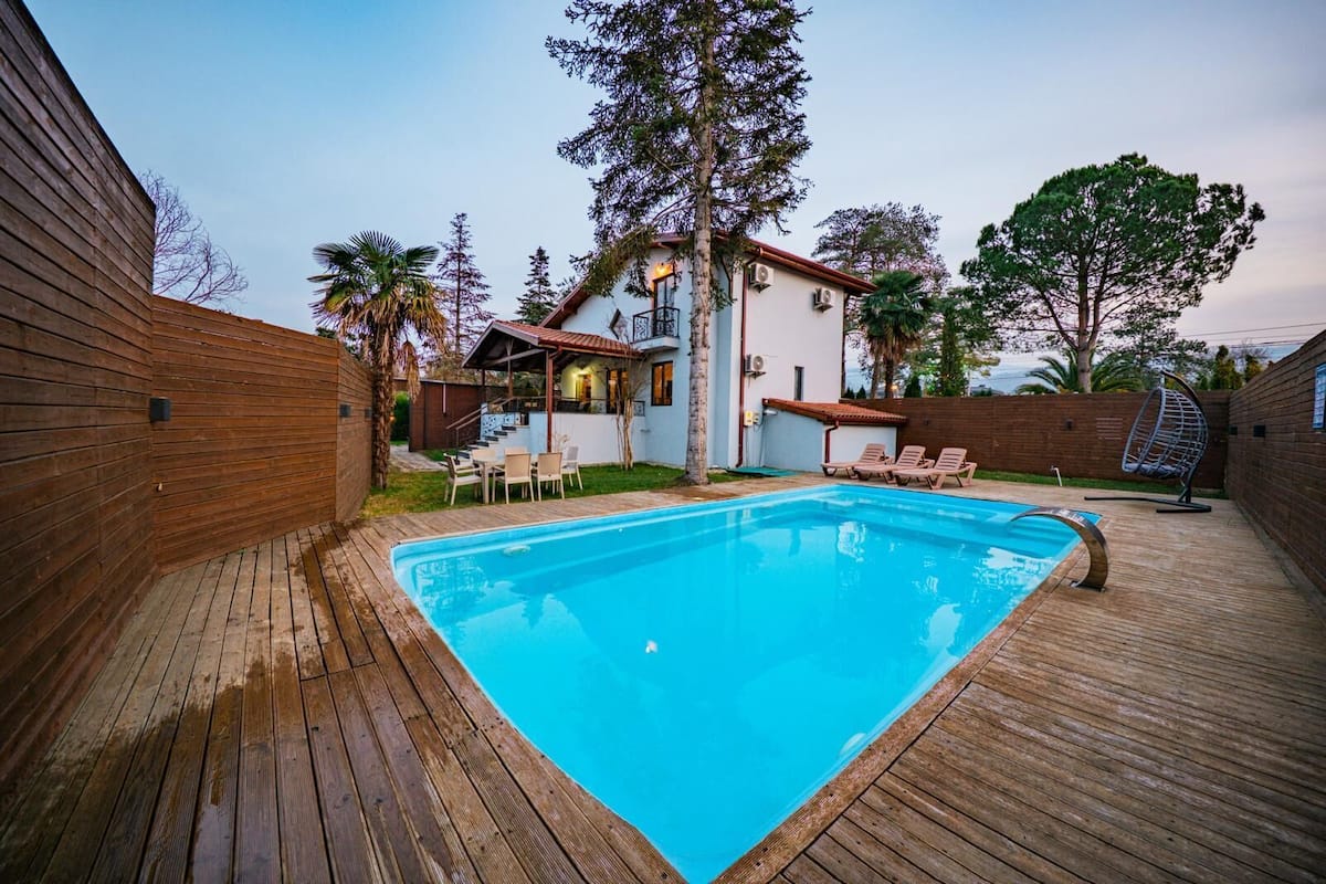 Una piscina rectangular con una terraza de madera alrededor, un pequeño patio con sillas y una hamaca colgante.