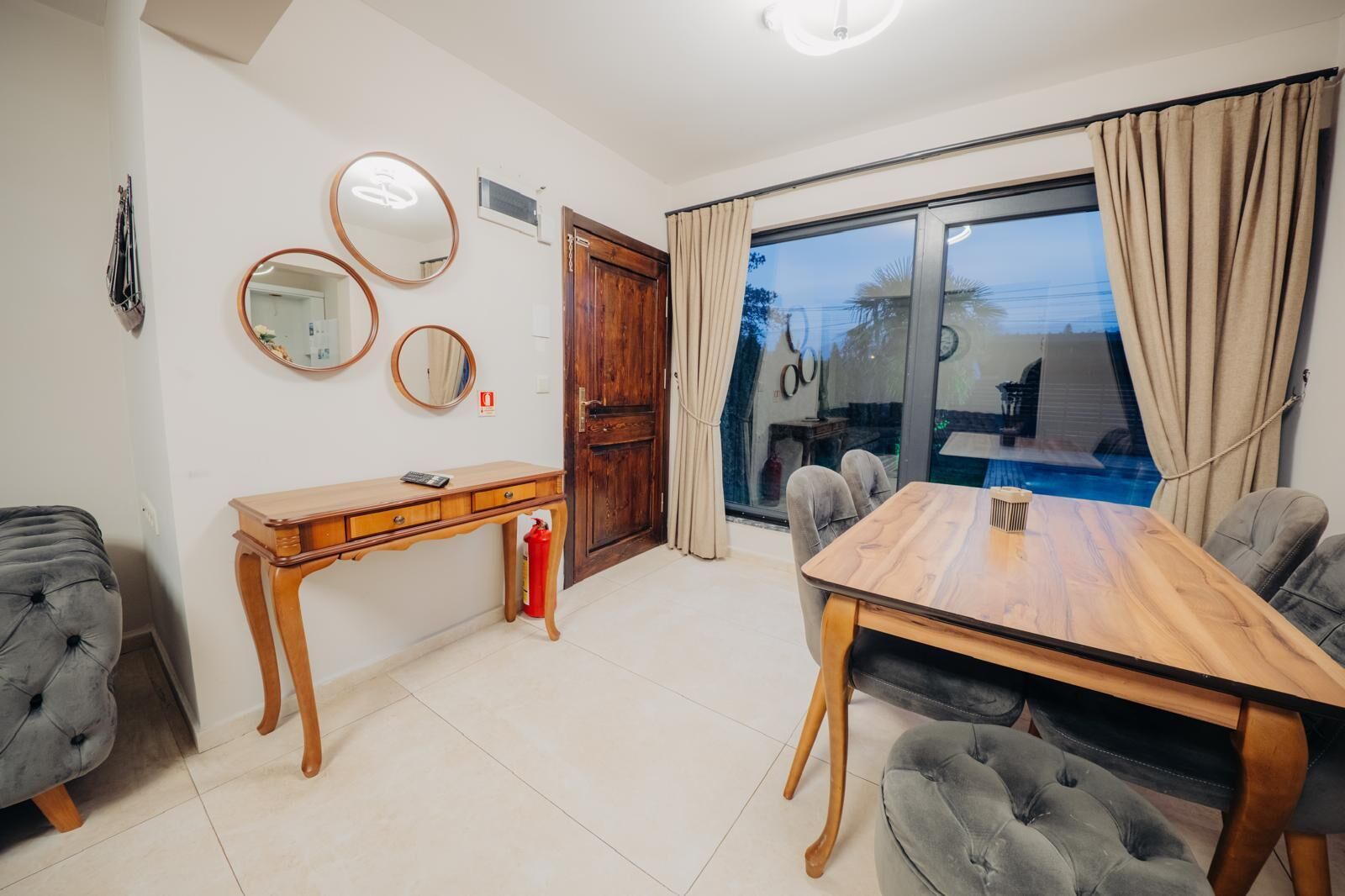 Una sala de estar moderna con una mesa de comedor de madera, sillas tapizadas de color gris, una mesita de noche de madera y un gran ventanal con cortinas.