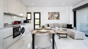 Cozinha moderna com armários brancos, área de jantar com mesa posta para dois e sala de estar com sofá e uma janela grande.
