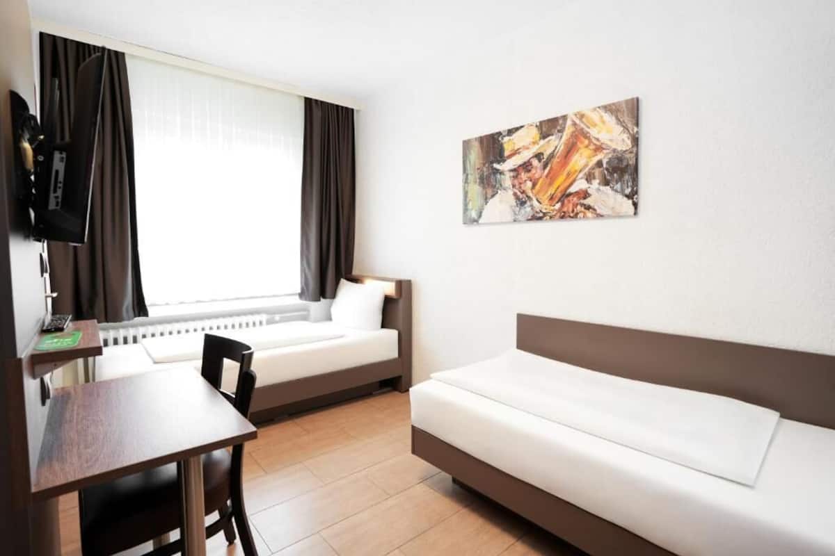 Ein Hotelzimmer mit Bett, Schreibtisch, Stuhl, einem Fenster mit Vorhängen, einem Bild an der Wand und einem Heizkörper.