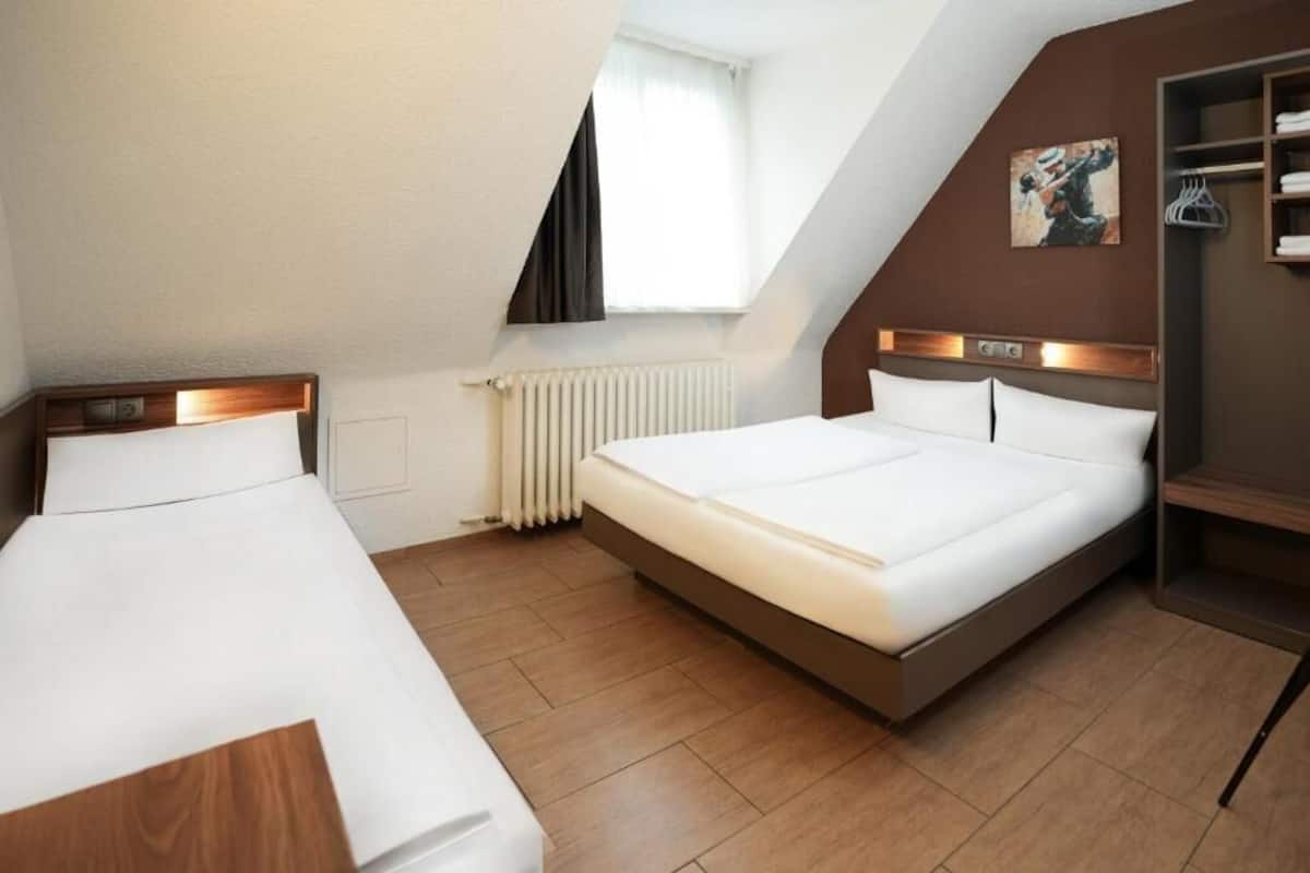 Ein Hotelzimmer mit einem großen Bett, einem kleineren Bett, einem Heizkörper, einem Fenster mit Vorhängen und einem Kleiderständer.