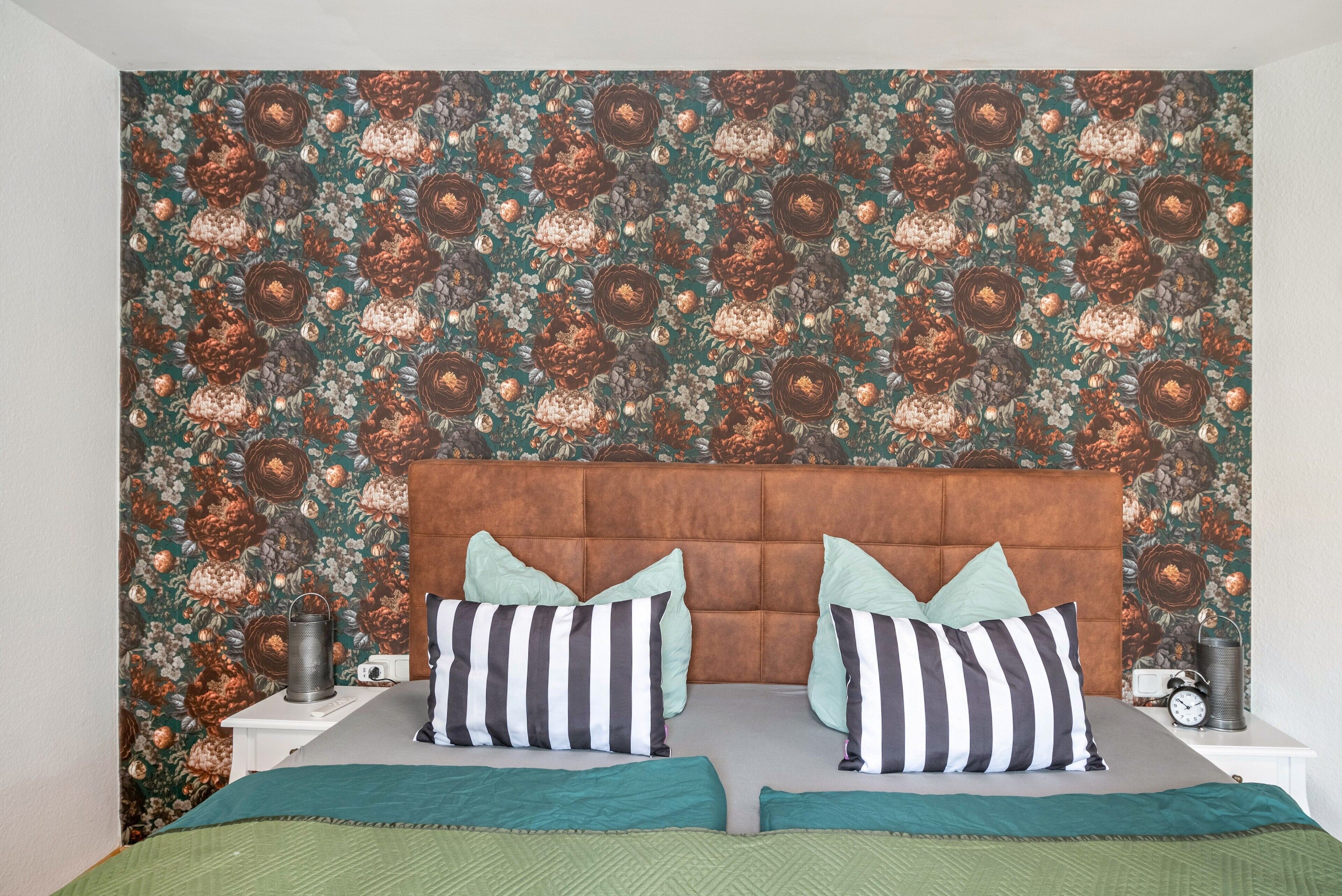 Ein Schlafzimmer mit gemustertem Wandpapier, einem gepolsterten Kopfteil, zwei Nachttischen mit Lampen und einem Bett mit gestreiften Kissen.