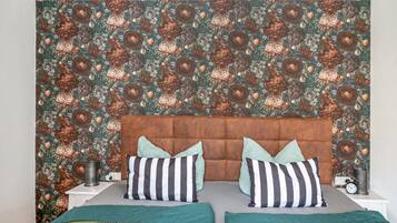 Ein Schlafzimmer mit gemustertem Wandpapier, einem gepolsterten Kopfteil, zwei Nachttischen mit Lampen und einem Bett mit gestreiften Kissen.
