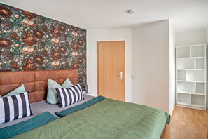 Ein Schlafzimmer mit ornamentalem Wandpapier, einer Holztür, einer weißen Regaleinheit und einer grünen Bettdecke.