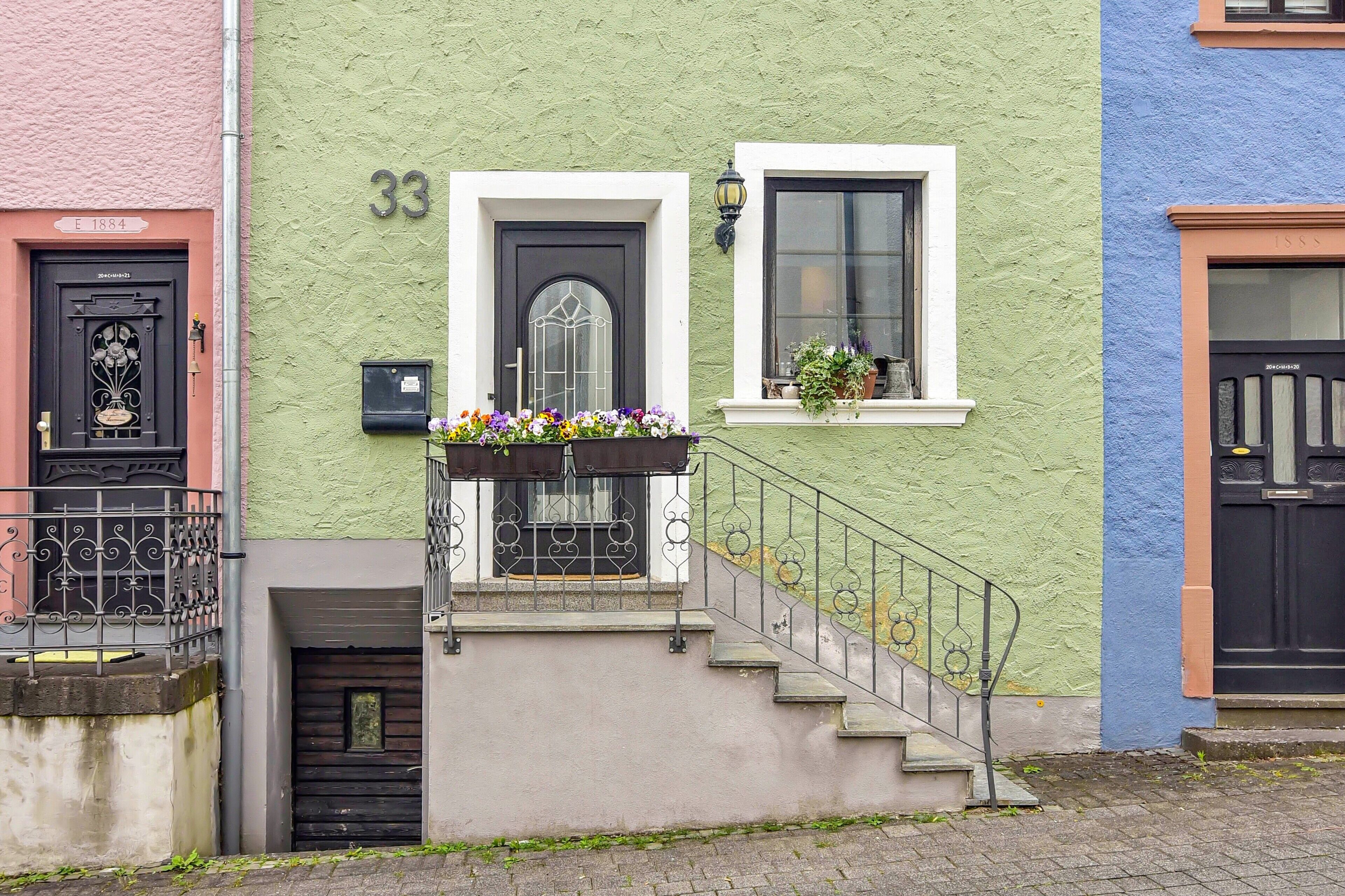 A building with a green facade, a black door with a decorative glass panel, a small balcony with flowers, and a white window with a black frame.