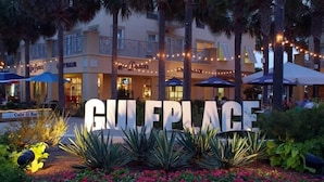 En hotellentrén med en stor skylt som visar texten ”GULF PLACE”, omgiven av tropiska växter och utomhusområden med sittplatser.