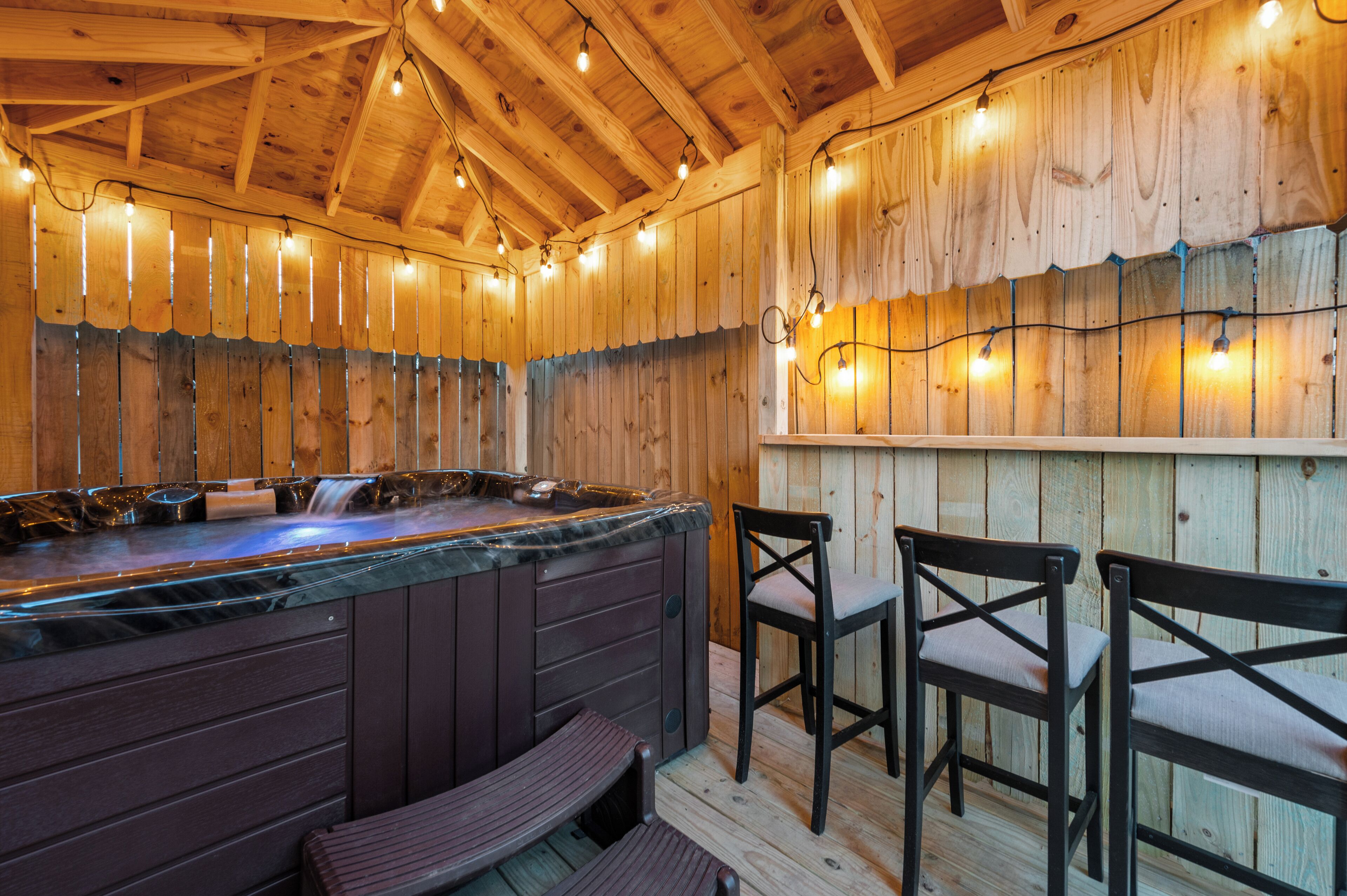 A wooden interior with a hot tub, wooden chairs, and string lights.