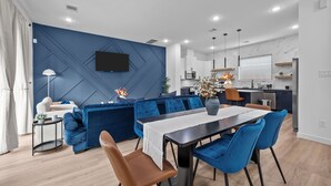 A modern living room with a blue accent wall, a dining area with blue chairs, and a kitchen in the background. - Central Houston 9BR Retreat – 2 Hot Tubs + Game Room – Sleeps 20+ (Houston)