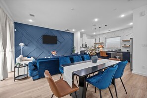 Ein modernes Wohnzimmer mit blauer Akzentwand, einem Essbereich mit blauen Stühlen und einer Küche im Hintergrund.