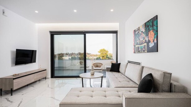 Uma sala de estar moderna com uma televisão grande, um sofá, uma mesa de centro e uma porta de vidro deslizante que dá para uma varanda.