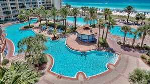 A large, multi-level swimming pool with palm trees and lounge areas, surrounded by a hotel building and a beach. - Shores 1613-King Bed-Beachfront Resort & Amenities (Panama City Beach)