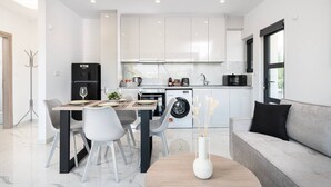 A modern kitchen with white cabinets, a dining area with a table and chairs, a washing machine, and a sofa. - Icon Luxury Apartments : A2 (Kos)