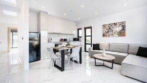 A modern kitchen with white cabinets, a dining area with a table and chairs, a gray sofa, and a colorful abstract painting on the wall. - Icon Luxury Apartments : A2 (Kos)