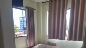 Een kamer met een bed, een stoel en een kleine tafel. Er is een raam met gordijnen, en een verwarming is aan de muur te zien.