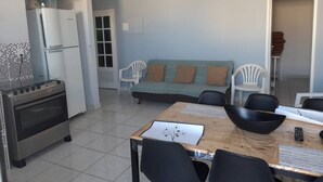 A compact kitchen with a stove, refrigerator, and tiled floor. There is a dining table with chairs and a sofa in the background. - Suite 303 beach walking distance (Anchieta)