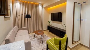 A modern living room with a sofa, a small table, a green armchair, a TV mounted on the wall, and a large mirror. - 7EVEN Suites (Lekki)