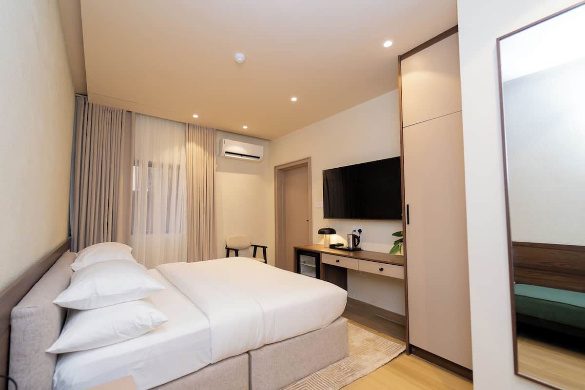 A modern hotel room with a large bed, a desk with a lamp, a flat-screen TV, and an air conditioning unit.