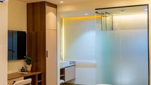A modern hotel room with a bed, a desk, a chair, a wardrobe, and a bathroom with a glass shower. - 7EVEN Suites (Lekki)