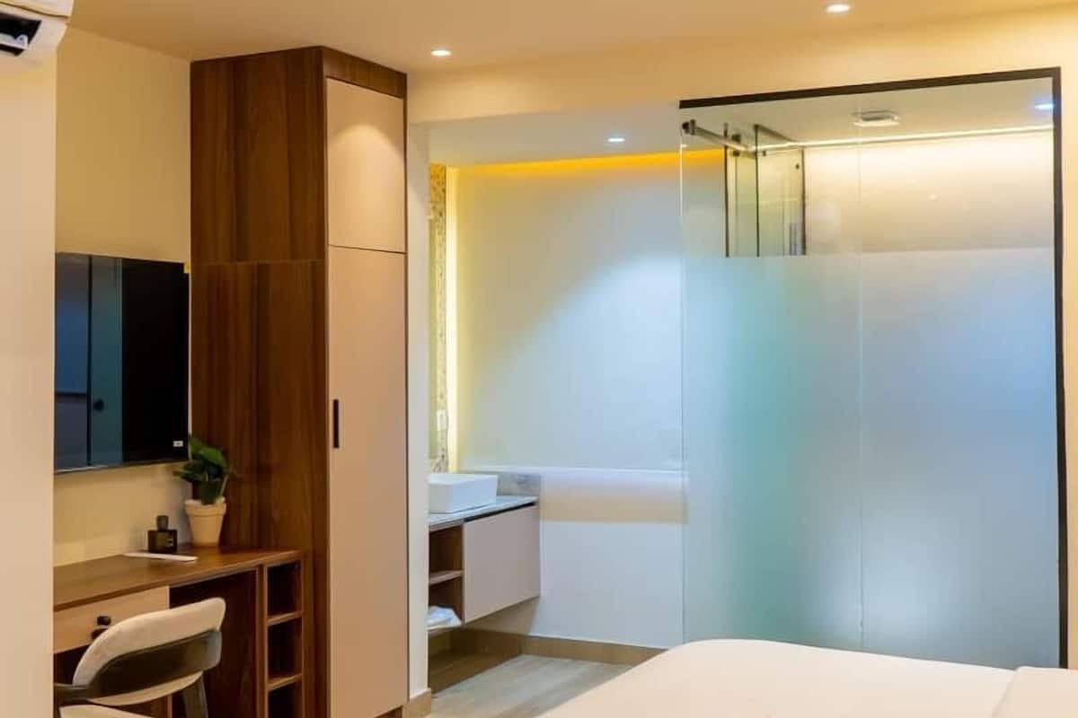 A modern hotel room with a bed, a desk, a chair, a wardrobe, and a bathroom with a glass shower.