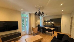 Ett modernt vardagsrum med en platt-TV, ett träbord, en svart soffa och en matplats med stolar. Köket syns i bakgrunden.