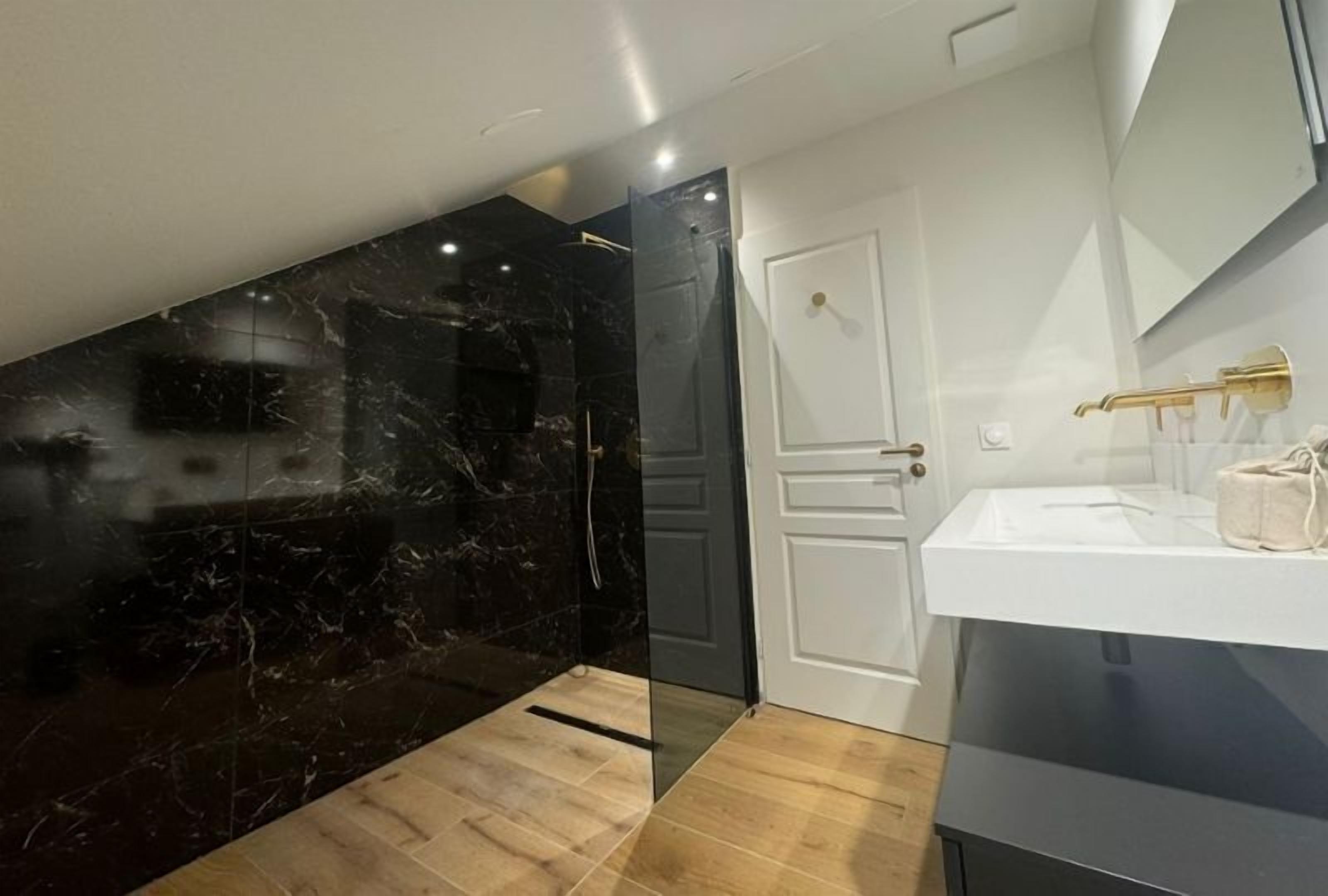 A modern bathroom with a black marble wall, a white door, and a white sink with gold fixtures.