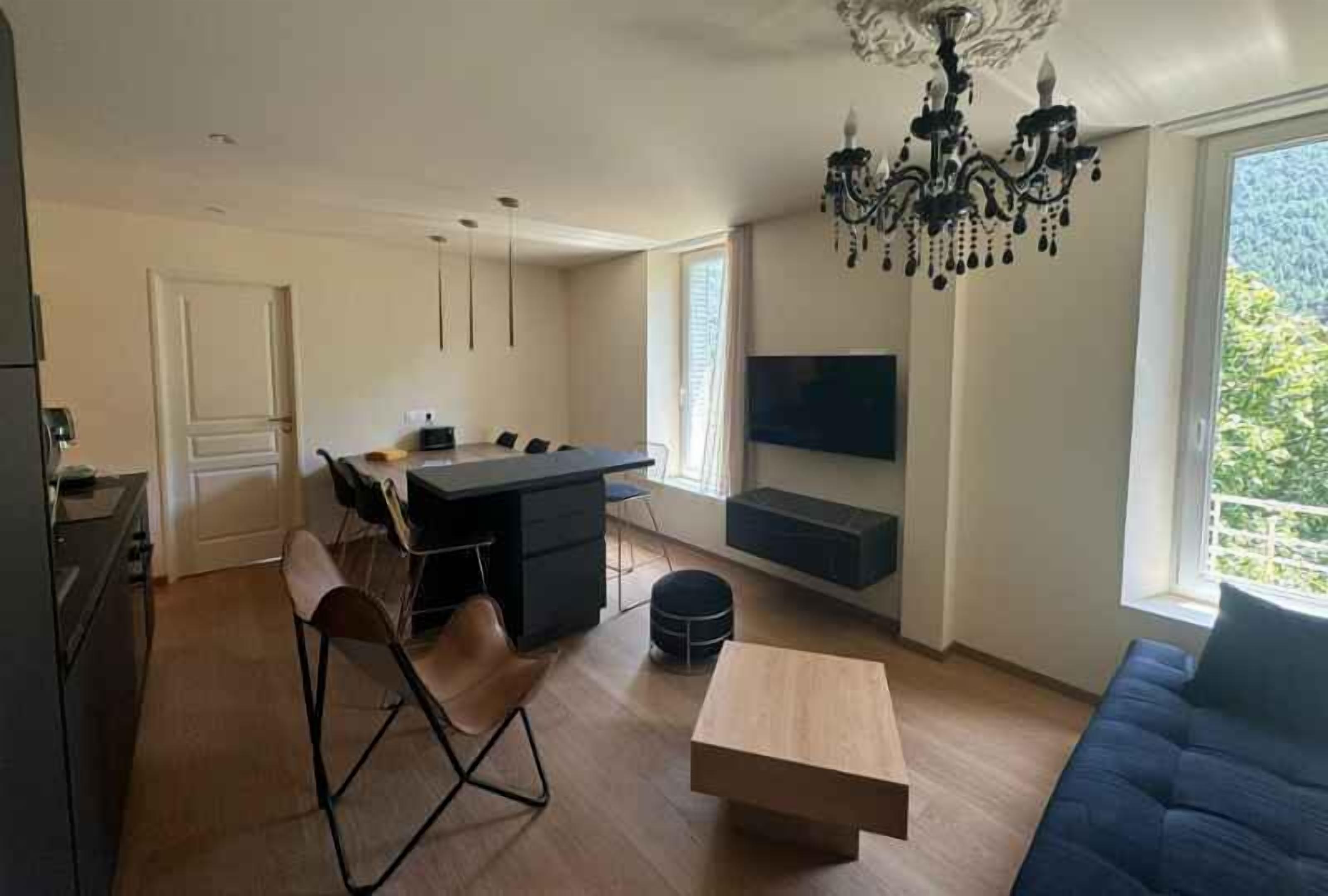 Een moderne woonkamer met een eethoek, een aan de muur bevestigde televisie en een groot raam dat voor natuurlijk licht zorgt.