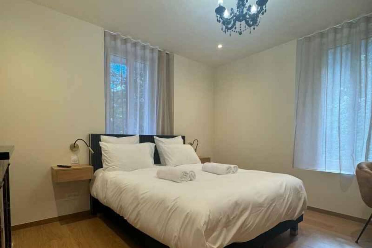 A neatly made bed with white linens, a bedside table with a lamp, and a chandelier.
