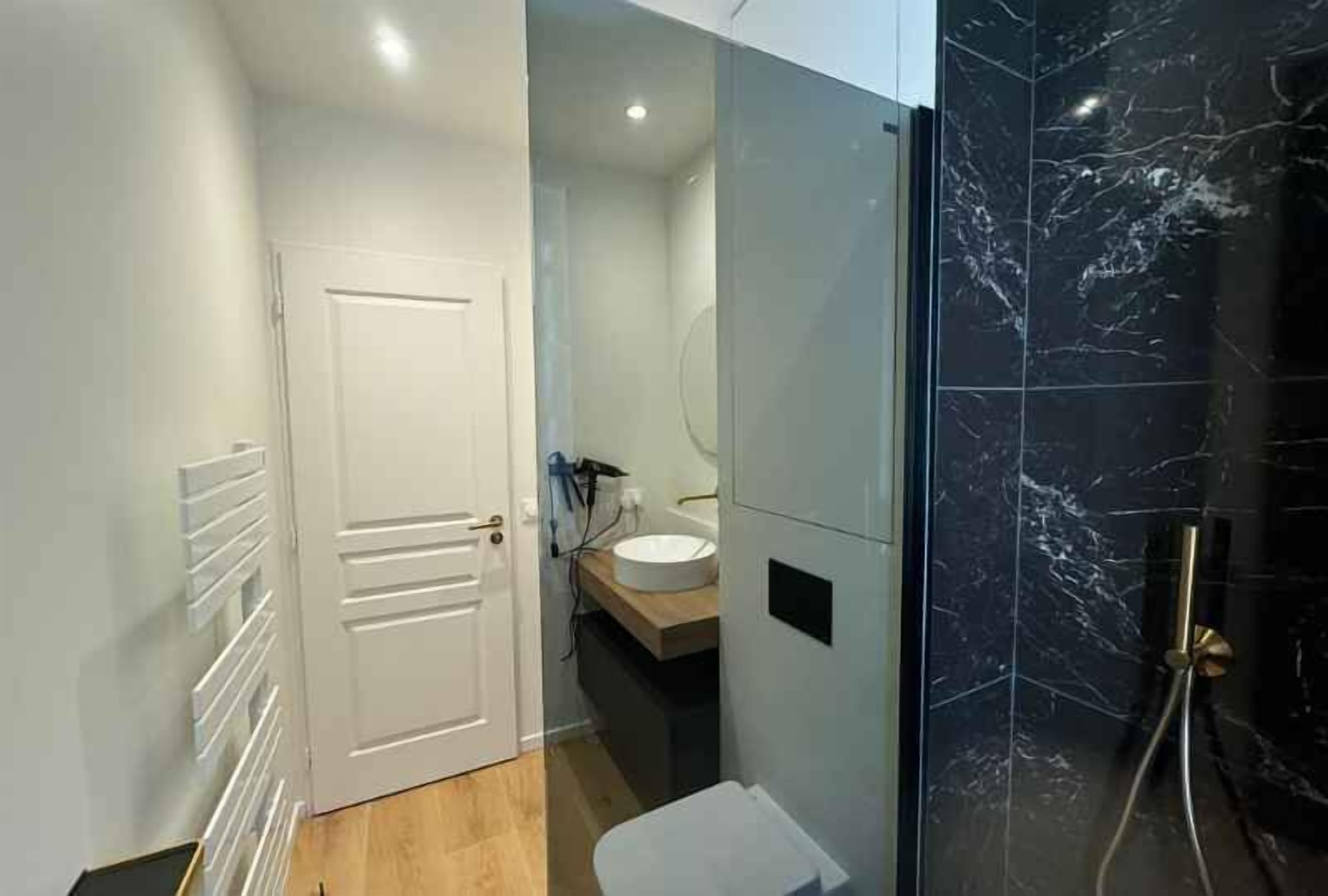 A modern bathroom with a white door, a wooden vanity, a white sink, and a black marble shower enclosure.
