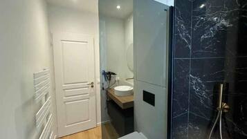 A modern bathroom with a white door, a wooden vanity, a white sink, and a black marble shower enclosure.