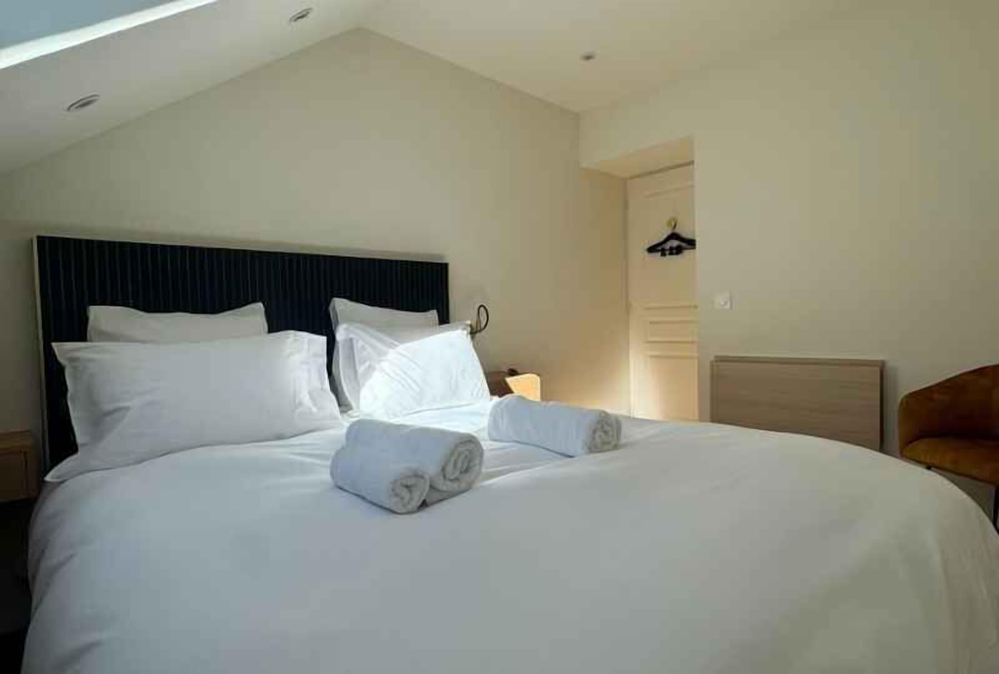 A neatly made bed with white linens and rolled towels, a dark headboard, and a wooden nightstand with a lamp.