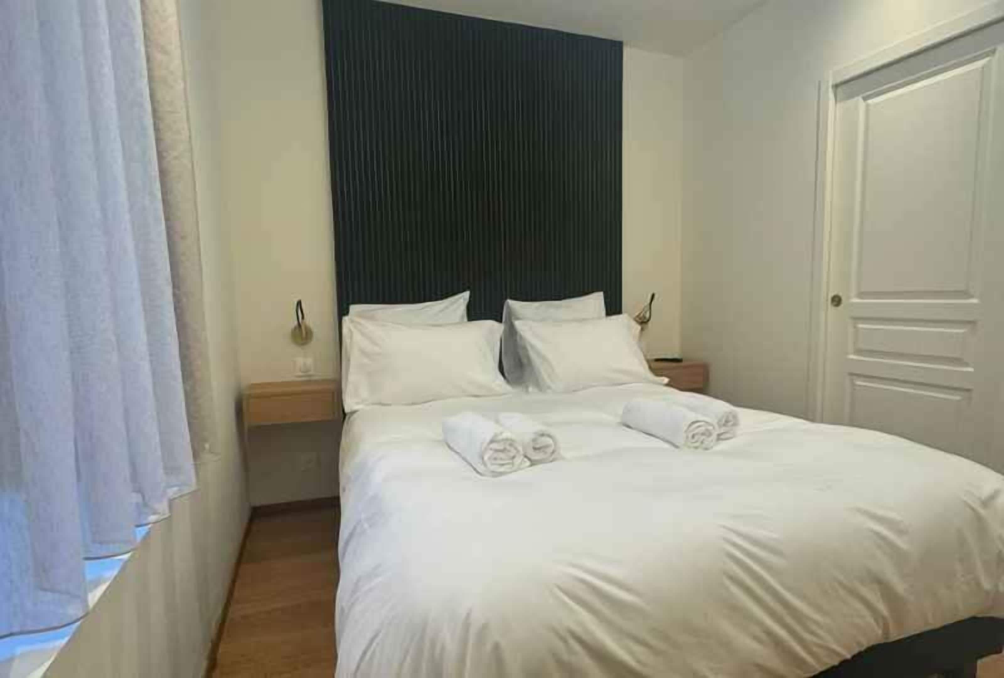 A neatly made bed with white linens and towels, a dark headboard, a bedside table with a lamp, and a closed door.