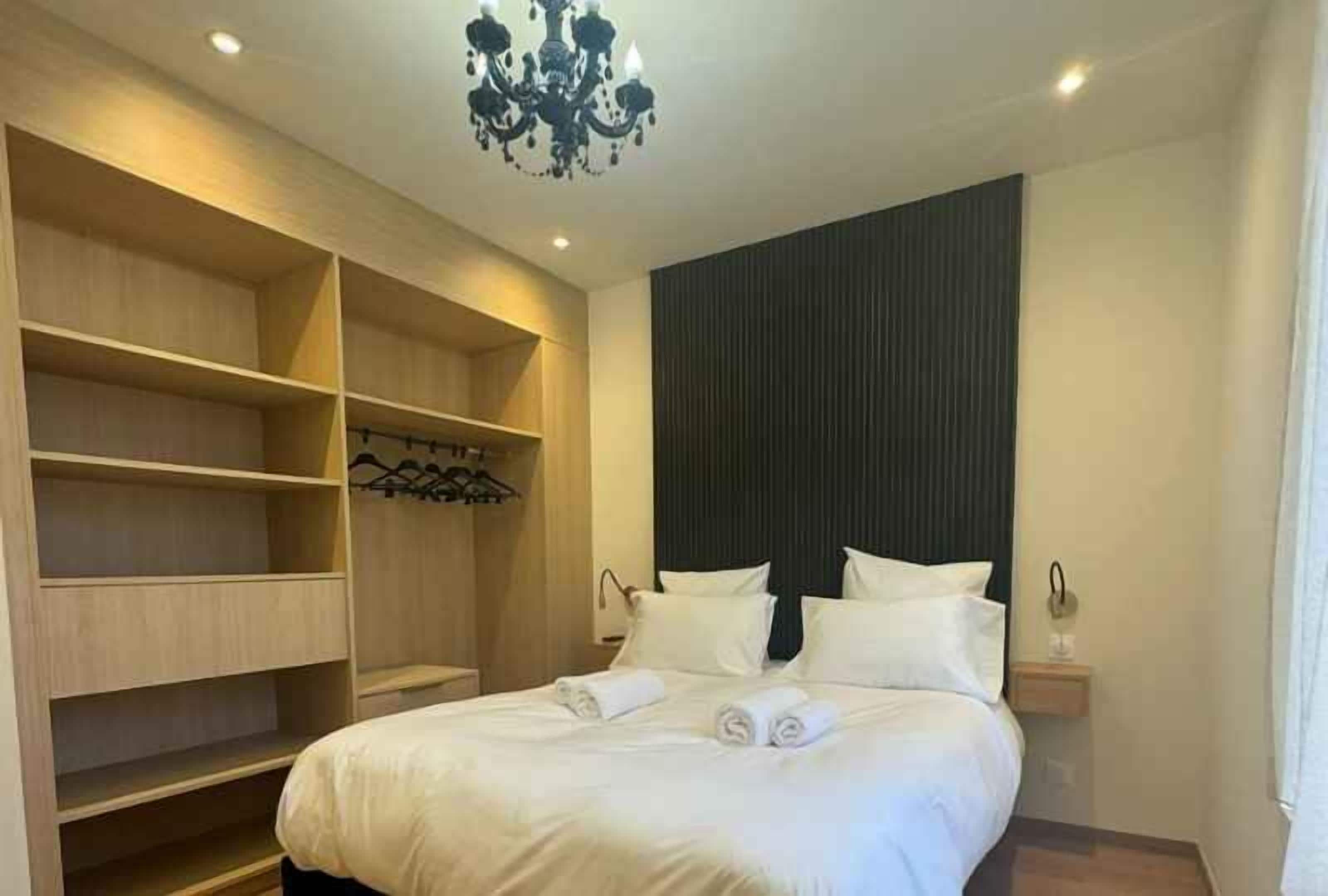 A hotel room with a large bed, built-in wooden shelving, a dark accent wall, and a chandelier.