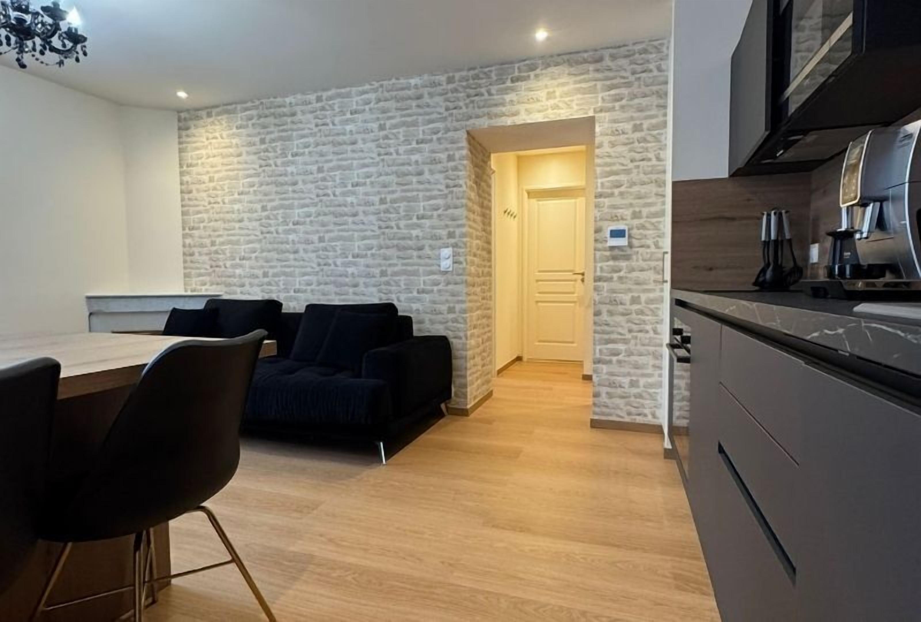 A modern kitchen with dark cabinets and a built-in oven. There is a dining area with a wooden table and black chairs. A stone wall separates the kitchen from the dining area.
