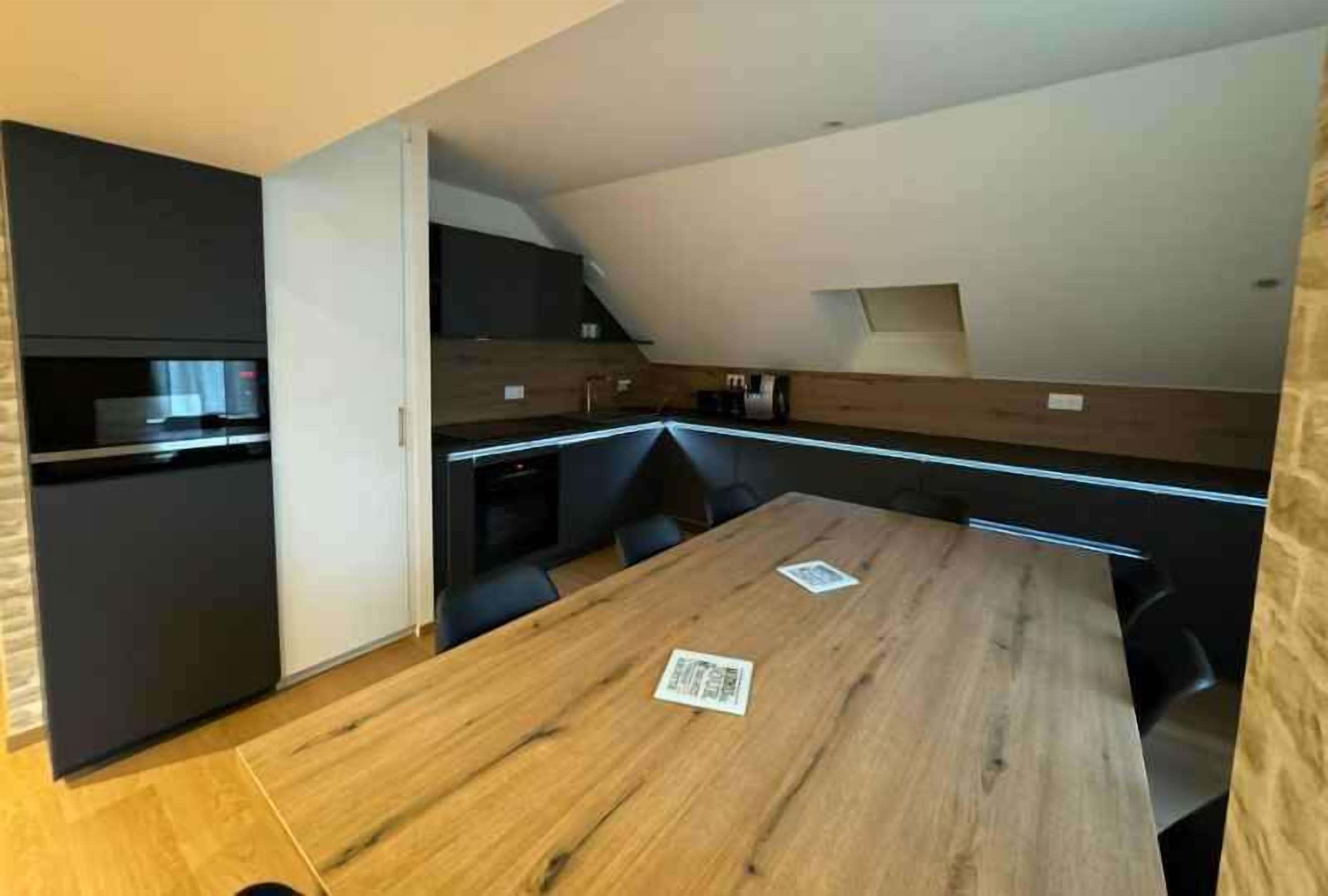 A modern kitchen with a wooden table, dark cabinetry, and a sloped ceiling.