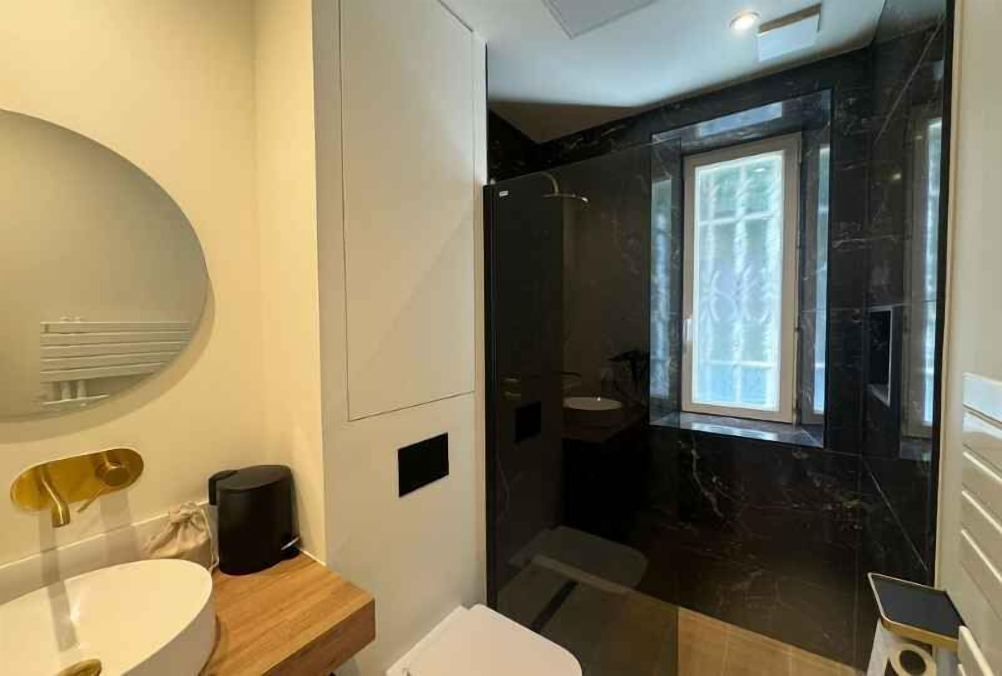 A modern bathroom with a white sink, a gold faucet, a wooden vanity, a black marble shower enclosure, and a large mirror.