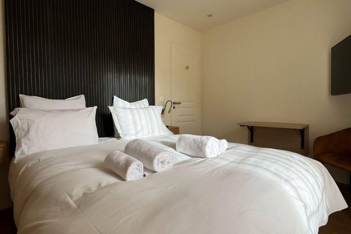 A neatly made bed with white linens and rolled towels, a wooden headboard, a small wooden table, and a brown chair in a hotel room.