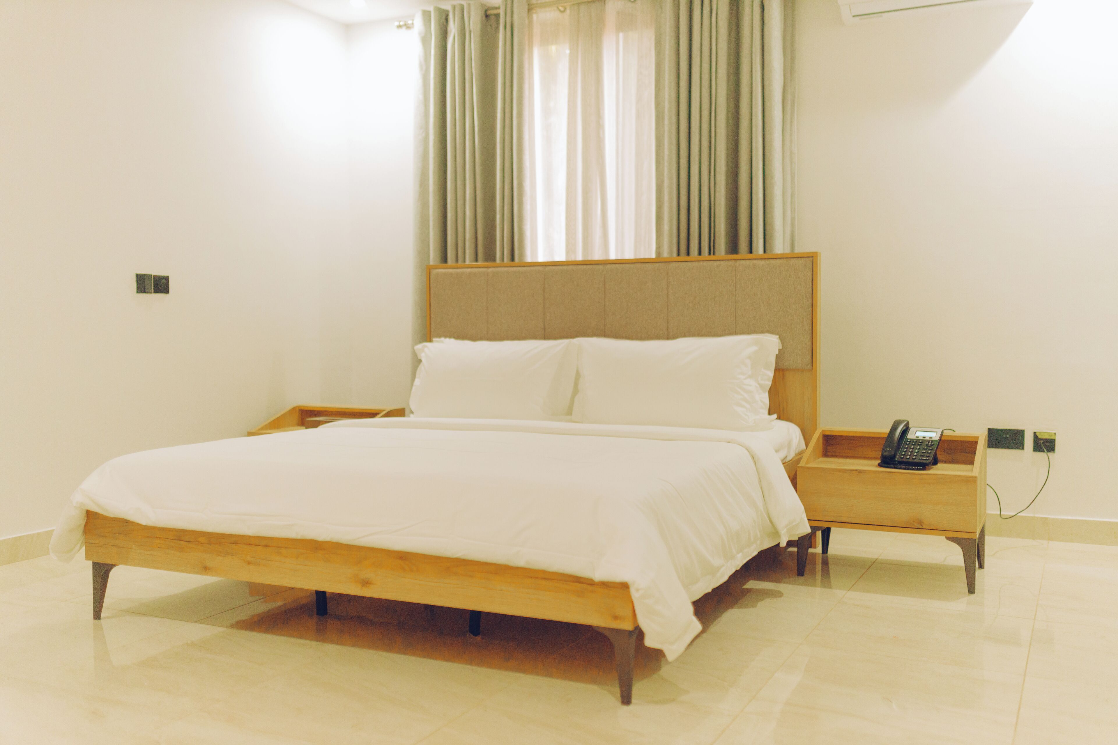 A neatly made bed with a wooden frame and white bedding, a bedside table with a telephone, and a window with curtains.