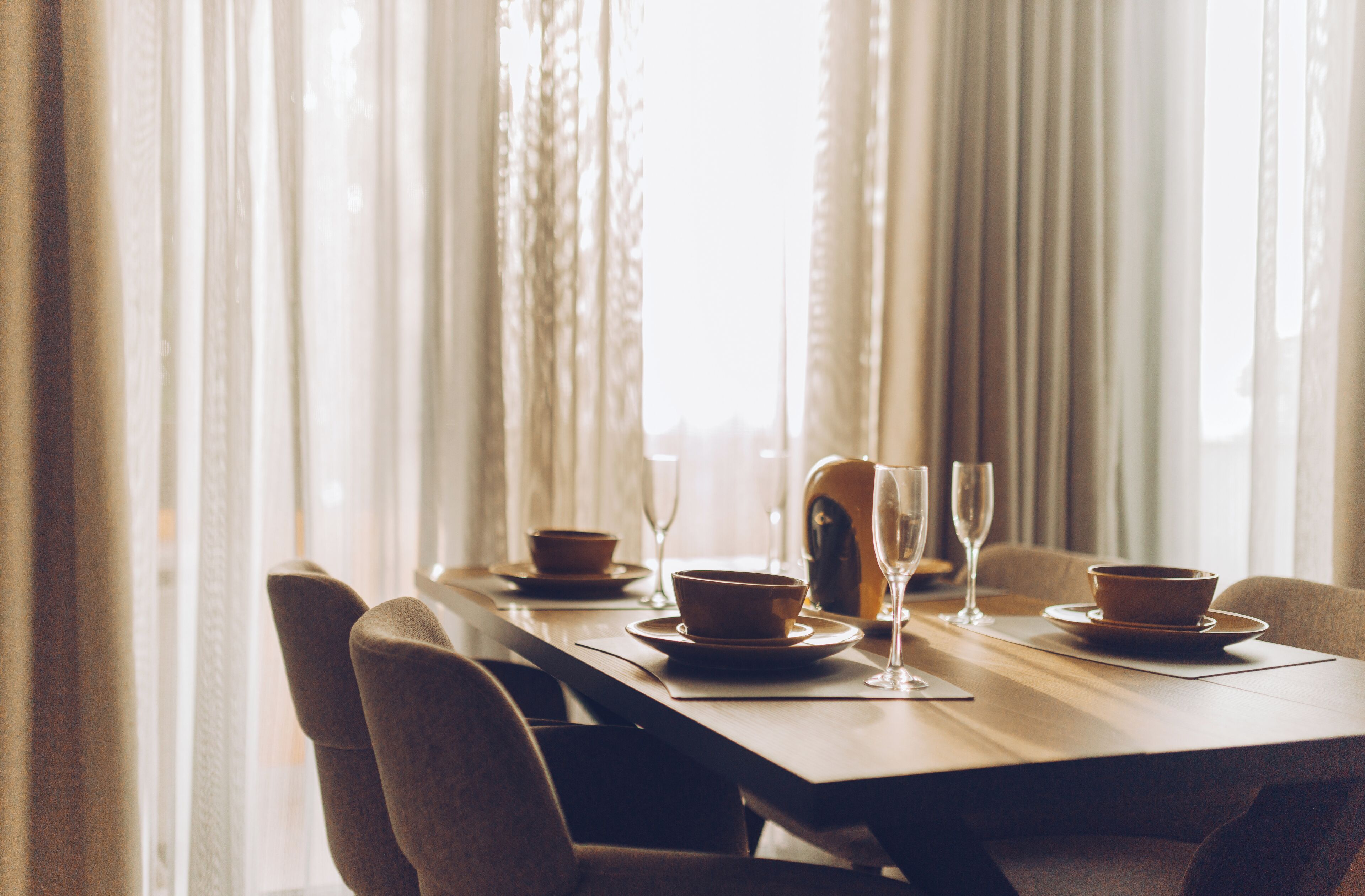 Una mesa de comedor dispuesta con cuencos y copas de vino, con vistas a cortinas y luz natural.