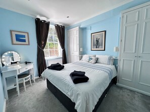 Ein Schlafzimmer mit Bett, Schminktisch mit Spiegel, Fenster mit Vorhängen und einem gerahmten Bild an der Wand.