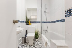 Un baño con lavamanos, inodoro y bañera blancos, un espejo sobre el lavamanos y una ducha con puerta de cristal.