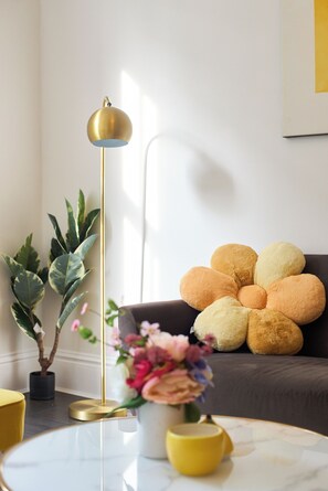 Ein modernes Wohnzimmer mit einem runden Glastisch, einer Stehlampe, einem Topfpflanzen und einem Sofa mit einem außergewöhnlichen Kissenmuster.